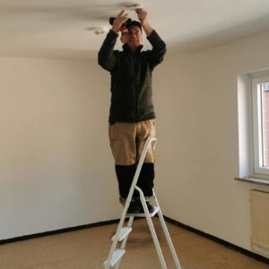 Photo by El Jundi Adult man on ladder fixing a ceiling light in an empty room with beige walls.