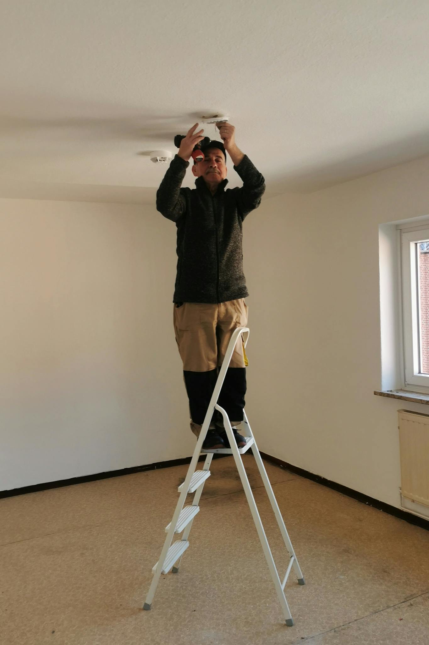 Adult man on ladder fixing a ceiling light in an empty room with beige walls.